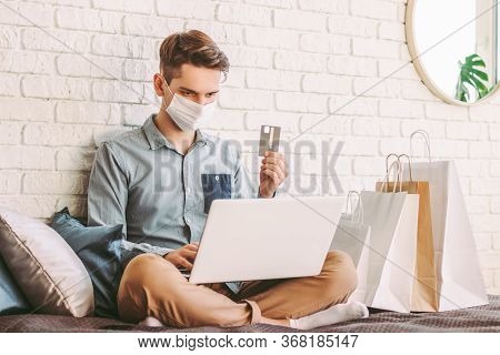 Young Male Shopper In Medical Face Mask Using Laptop On Couch For Online Shopping. Hipster Man In Pr