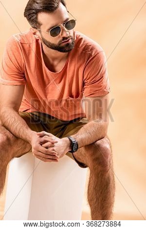 Handsome Man In Shorts, Summer T-shirt And Sunglasses Sitting On White Cube On Beige