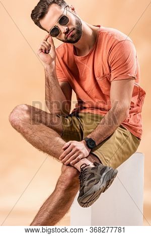 Young Man In Shorts And Summer T-shirt Holding Sunglasses Sitting On White Cube On Beige