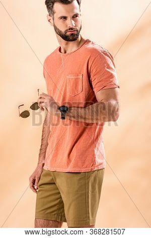 Handsome Stylish Man Posing In Shorts And Summer T-shirt Holding Sunglasses On Beige