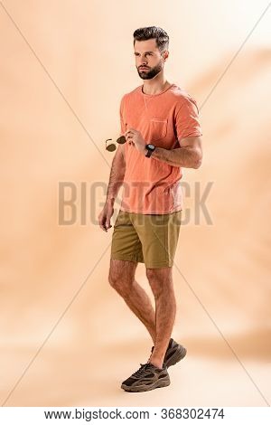 Stylish Handsome Man Posing In Shorts And Summer T-shirt Holding Sunglasses On Beige