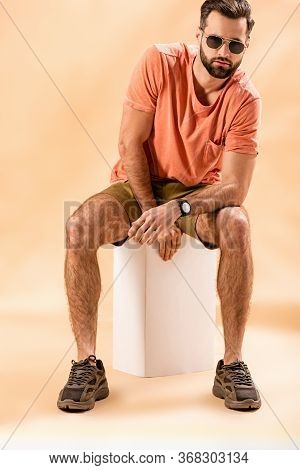 Fashionable Young Man In Shorts, Summer T-shirt And Sunglasses Sitting On White Cube On Beige