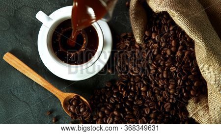 Pouring a cup of hot coffee, high angle of view. Still life scene made of pile of roasted coffee beans.