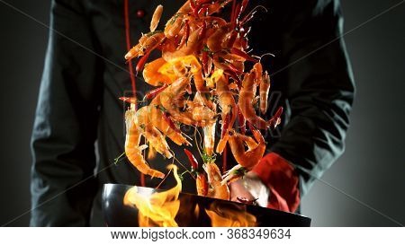 Closeup of chef throwing prawns from wok pan in fire. Fresh asian food preparation on dark background.