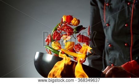 Closeup of chef throwing chilli peppers mix from wok pan in fire. Fresh asian food preparation on dark background.