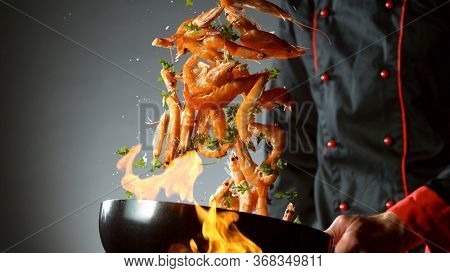 Closeup of chef throwing prawns from wok pan in fire. Fresh asian food preparation on dark background.