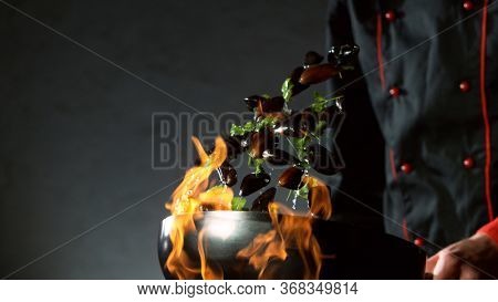 Closeup of chef throwing mussels from wok pan in fire. Fresh asian food preparation on dark background.