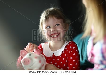 Portrait Of Smiling Beautiful Little Girl Holding Piggy Bank On Couch At Home. Pretty Blonde Kid Sav
