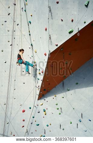 Athletic Woman Climber In Bright Blue Pants Swinging On Overhanging Artificia Rock Wall With Rope.