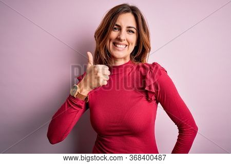 Young beautiful brunette woman wearing casual sweater stnding over pink background doing happy thumbs up gesture with hand. Approving expression looking at the camera showing success.