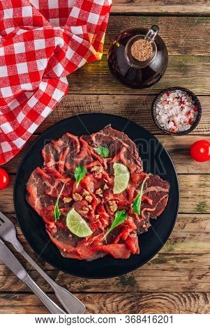 Black Plate With Carpaccio Of Beef With Baby Arugul And Lime On Rustic Wooden Background. Top View. 
