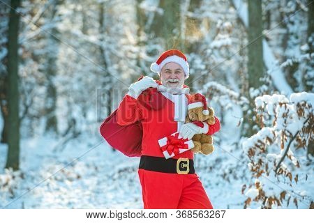 Real Santa Claus In Red Cap Pulling Large Red Gift Sack. Santa Carrying Sack Of Gifts Against Snow S