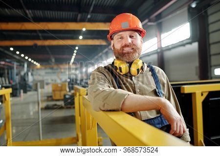 Portrait of positive male worker in orange hardhat leaning on metal railings in modern factory shop