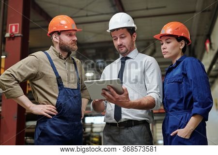 Industrial engineer in formalwear and hardhat discussing machine part with workers using 3D sketch on tablet