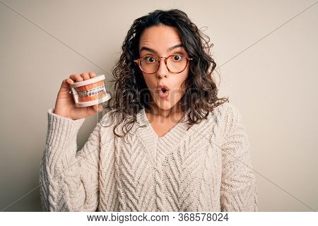 Young beautiful woman with curly hair holding plastic teeth with dental baces scared in shock with a surprise face, afraid and excited with fear expression