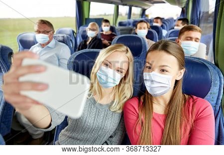 transport, tourism, road trip and people concept - young women or friends wearing face protective medical mask for protection from virus disease in travel bus taking selfie by smartphone