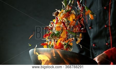 Closeup of chef throwing vegetable mix from wok pan in fire. Fresh asian food preparation on dark background.