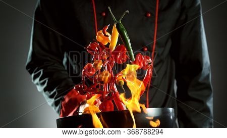 Closeup of chef throwing chilli peppers mix from wok pan in fire. Fresh asian food preparation on dark background.
