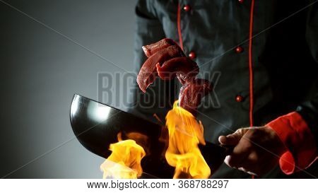 Closeup of chef throwing raw beef steaks from wok pan in fire. Fresh asian food preparation on dark background.