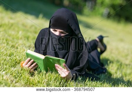 Islamic Woman Reads A Book On A Lawn In A Park. Muslim Student Girl Preparing For An Exam Reads A Te
