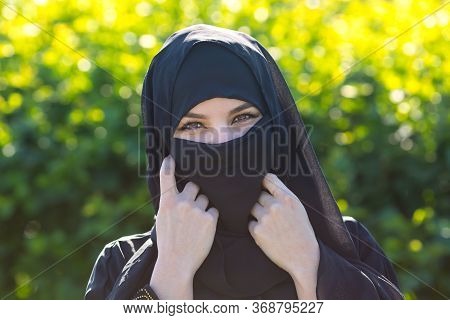 Beautiful Muslim Woman On A Background Of Green Leaves. Islamic Woman In Oriental Clothes Stands Aga
