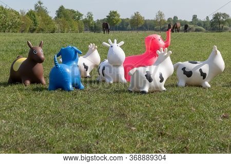 A Herd Of Inflatable Hopper Toys In The Field. Green With Yellow Flowers Pasture, Trees In The Backg