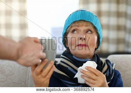 Elderly Woman In Scarf And Hat Sits On Sofa Home. Elderly Lady Takes Cup Tea. Weak Immune System In 