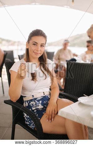 Beautiful Woman Drink Coffe In Cafe Outside. Background Of Sea. Happy Young Woman Sits On Terrace Wi