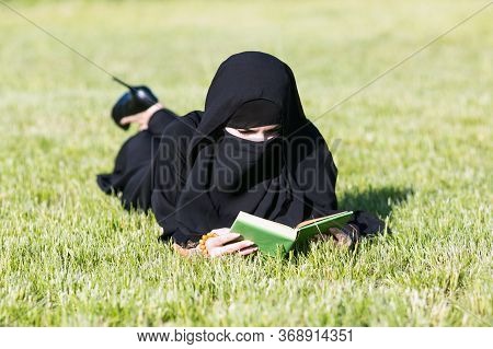 Islamic Woman Reads A Book On A Lawn In A Park. Muslim Student Girl Preparing For An Exam Reads Text