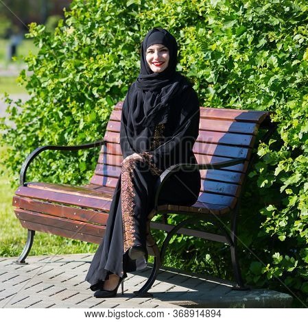 A Beautiful Young Muslim Woman Is Sitting On A Bench. Islamic Girl Smiles Broadly, Resting On A Park