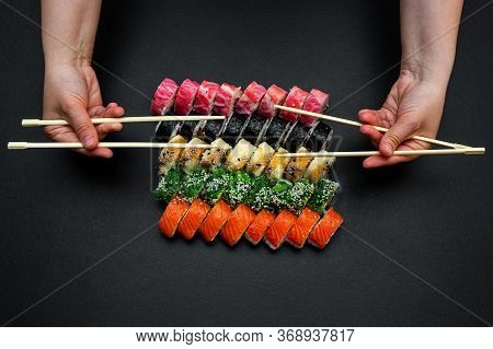 Big Sushi Set On A Black Plate Background. A Female Hand Holds Two Paar Chopsticks.