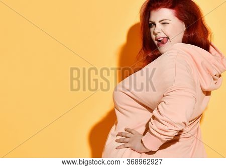 Side Shot Of Joking Positive Chubby Young Female Standing With Hands On Hips, Giving A Wink And Loll