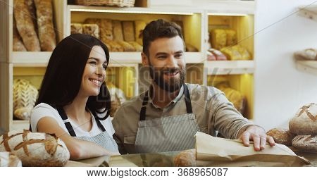 Charming Female Vendor In The Bakery Shop Selling Baguettes While Her Male Co-worker Helping Her. At