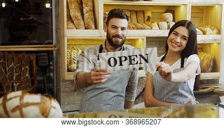 Portrait Of The Male And Female Caucasian Smiled Bakers Holding Together A Signboard Open And Posing