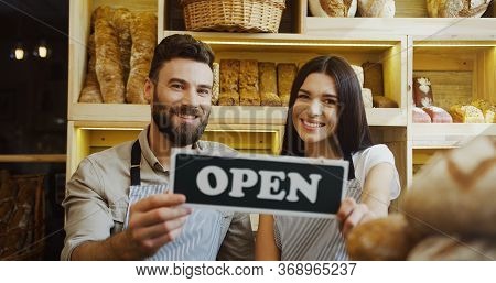 Portrait Of The Male And Female Caucasian Smiled Bakers Holding Together A Signboard Open And Posing