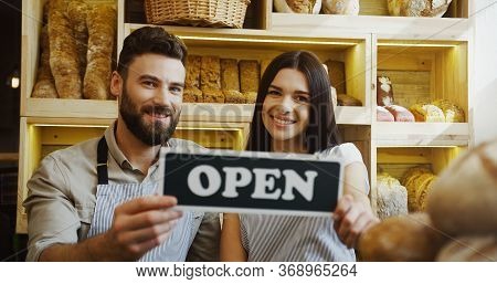 Portrait Of The Male And Female Caucasian Smiled Bakers Holding Together A Signboard Open And Posing