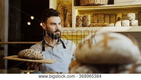 Portrait Shot Of The Caucasian Young Male Baker Looking At The Side While Holding A Tray With Bread 
