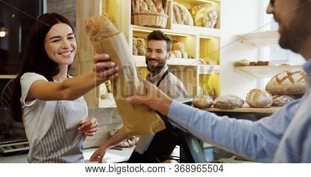 Beautiful Woman, Seller In The Bakery Shop, Selling Baguettes To The Man While Her Male Handsome Co-