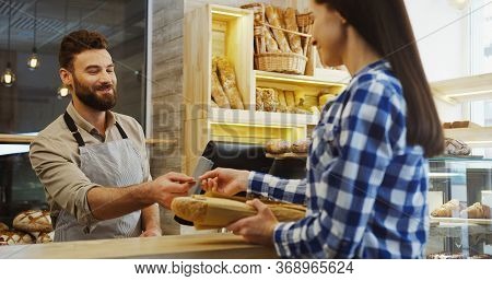 Handsome Man, Bakery Vendor Selling Baguettes To The Young Woman And She Paying With A Credit Card. 