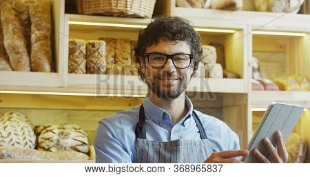 Attractive Male Bread Vendor In Glasses Scrolling And Taping On The Tablet Device While Standing At 