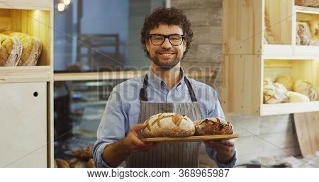 Portrait Shot Of The Attractive Young Man, Bakery Vendor, Holding A Tray With Fresh Bread And Lookin