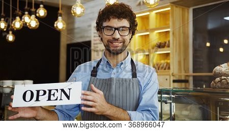 Male Caucasian Smiled Bakery Vendor Posing With A Signboard Open And Smiling In The Shop.