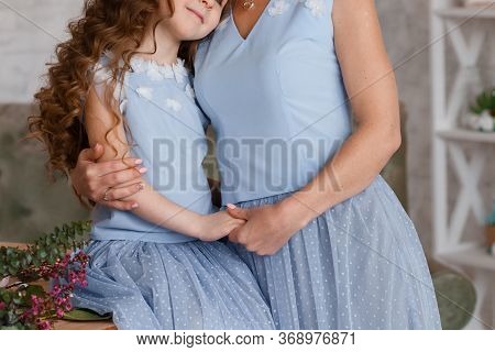 A Young Woman Stands Near A Wooden Table And Holds Her Daughter's Hand. They Are Dressed In Blue Dre