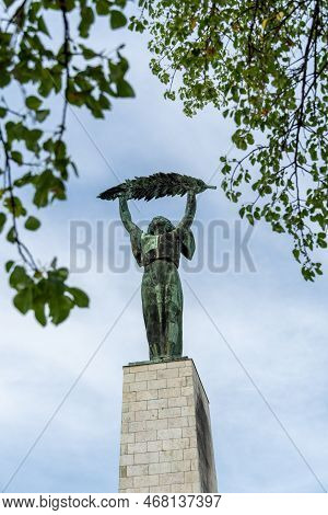 The Historic Liberty Statue Upon Gellert Hill In Front Of A Cloudy Sky And Green Leaves In Budapest,