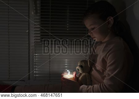 A Girl In A Dark Room Sits Near The Window And Holds A Candle And A Teddy Bear In Her Hands. Power O