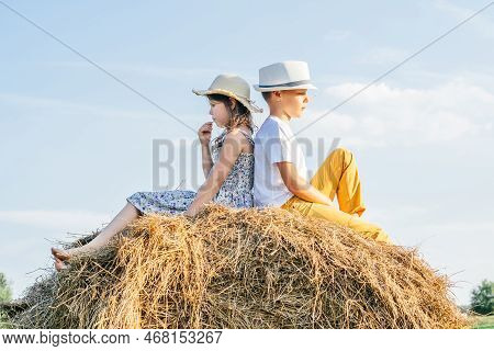 Thoughtful Little Girl And Boy Sitting Back To Back At Top Of Haystack. Friends Resting On Hayrick, 