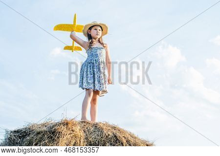 Serious Little Girl In Hat Stand At Top Of Haystack And Launch Yellow Toy Airplane. Child On Hayrick