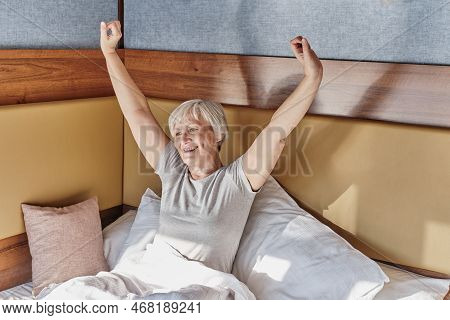 Happy Senior Smiling Woman With Short Gray Hair Stretching Oneself After Sleeping In Bed, Enjoying S
