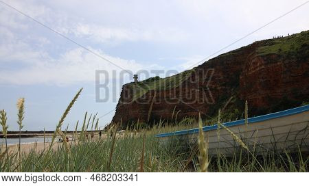 Low Angle View Over Bolata Coast Cliff And Lighthouse With A Blue Boat And The Beach In Foreground, 