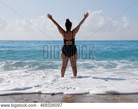 A Curvaceous Woman In A Black Bathing Suit Stands In The Sea, Raising Her Hands